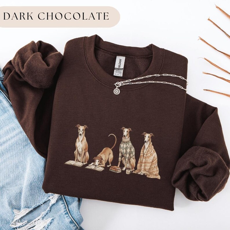 May include: Dark chocolate-colored sweatshirt featuring a graphic of four greyhounds, each with unique attire and poses. One is reading a book, another is wearing a sweater, and another is draped in a blanket. The text "DARK CHOCOLATE" is at the top.