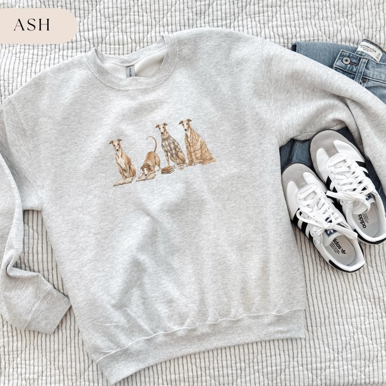 May include: A light gray sweatshirt with a crew neck featuring a watercolor illustration of four dogs. The word "ASH" is in a light beige rectangle in the upper left corner. The sweatshirt is paired with blue jeans and white sneakers.
