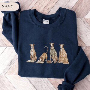 May include: Navy blue sweatshirt featuring a whimsical illustration of four greyhounds, each depicted in different poses with books and cozy attire. The word "NAVY" is in a cream-colored oval in the upper left corner. The sweatshirt is made of a soft material.