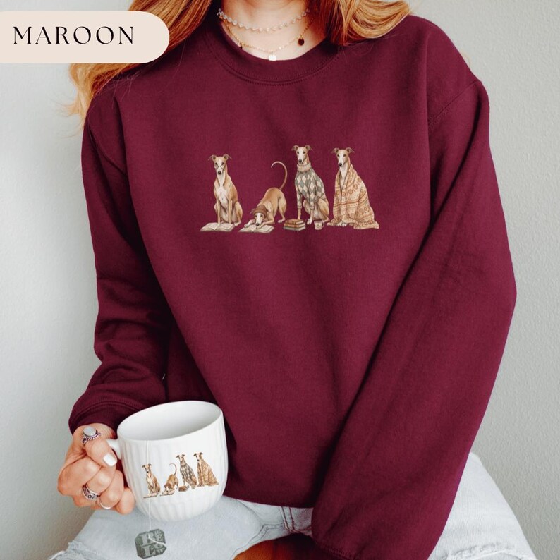May include: Maroon sweatshirt featuring a graphic of four greyhounds in various poses. The dogs are depicted in a watercolor style, with one lying down and others sitting. The word "MAROON" is in a cream-colored rectangle in the upper left corner.