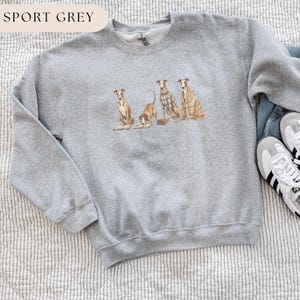 May include: Sport grey sweatshirt featuring a watercolor illustration of four greyhounds. The dogs are in various poses, with one lying down and others sitting. The sweatshirt has a crew neck and long sleeves. The text "SPORT GREY" is at the top.