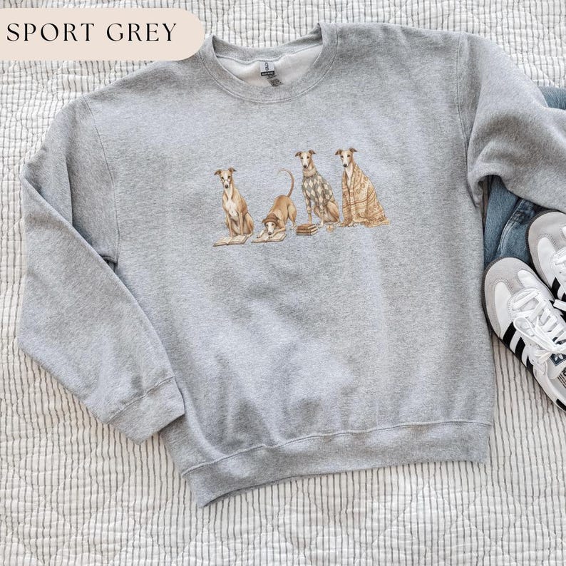 May include: Sport grey sweatshirt featuring a watercolor illustration of four greyhounds. The dogs are in various poses, with one lying down and others sitting. The sweatshirt has a crew neck and long sleeves. The text "SPORT GREY" is at the top.