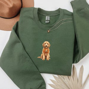 May include: An olive green crewneck sweatshirt featuring an embroidered graphic of a golden-colored dog holding a small orange jack-o'-lantern pumpkin in its mouth. The sweatshirt has long sleeves and a ribbed collar, cuffs, and hem.