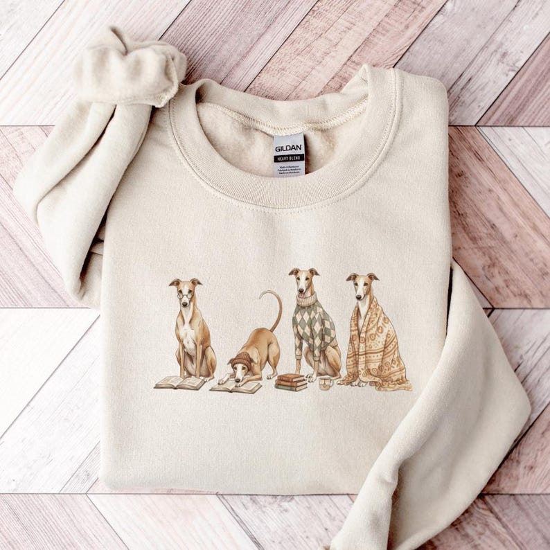 May include: Cream-colored sweatshirt featuring a watercolor illustration of four greyhounds. Each dog is depicted in a different pose, some with books, and one wearing a sweater. The sweatshirt has a crew neck and long sleeves.