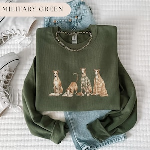 May include: Military green sweatshirt featuring a graphic of four greyhounds, each with unique attire, including a sweater and a blanket. The text "MILITARY GREEN" is at the top. A gold chain necklace is draped on the sweatshirt.