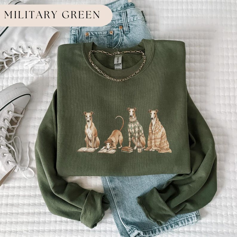 May include: Military green sweatshirt featuring a graphic of four greyhounds, each with unique attire, including a sweater and a blanket. The text "MILITARY GREEN" is at the top. A gold chain necklace is draped on the sweatshirt.