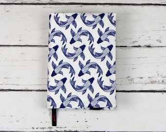 Puede incluir: Una cubierta de cuaderno blanca con un patrón de peces koi azules y blancos. La cubierta tiene un marcapáginas de cinta negra.