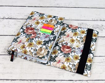 May include: A white fabric notebook cover with a floral pattern in shades of pink, yellow, and green. The cover has a black elastic band closure and a red bookmark.