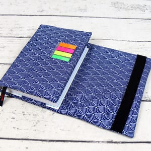 May include: A blue notebook with a white wave pattern, open to reveal the pages. A black elastic band holds the cover closed. Colorful sticky notes are attached to the top of the notebook. A red bookmark is visible.