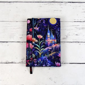 Pode incluir: Uma capa de livro azul escuro com uma ilustração colorida de um castelo em uma floresta à noite. O castelo está iluminado com luzes amarelas e há uma lua cheia no céu. A floresta está cheia de flores e plantas em tons de rosa, roxo e verde.