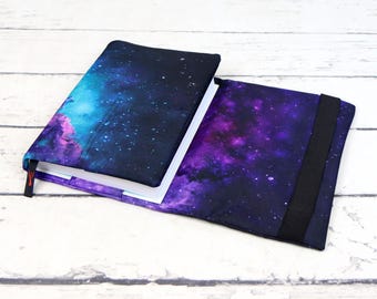 Puede incluir: Una funda para libro con estampado de galaxia y una correa negra. La funda está hecha de tela y tiene una combinación de colores azul, morado y negro.