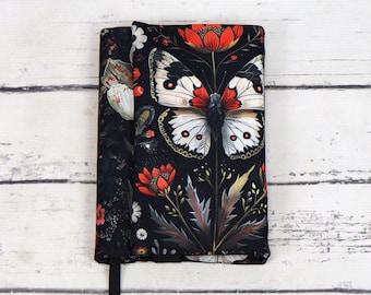Puede incluir: Funda de libro de tela negra con una mariposa blanca y flores rojas. La mariposa tiene marcas negras y manchas blancas. Las flores son rojas con centros amarillos.