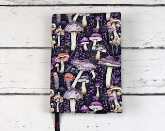 Puede incluir: Una cubierta de cuaderno negra con un patrón de setas coloridas. Las setas son moradas, rosas y blancas con tallos marrones. El fondo es morado oscuro.