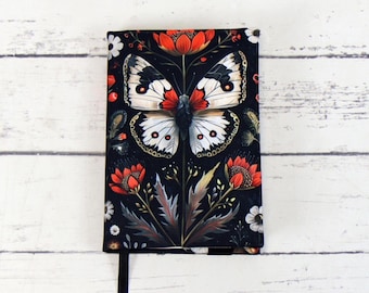 Puede incluir: Cubierta de cuaderno negra con una mariposa blanca con detalles rojos sobre un diseño floral con detalles rojos, blancos y dorados.