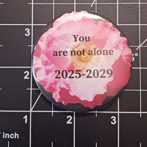 May include: A pink and white flower button with the text "You are not alone 2025-2029" on it.