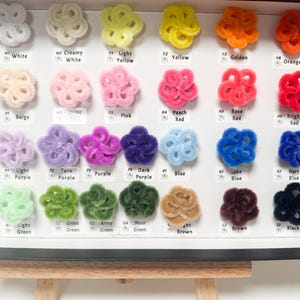 May include: A display of colorful, fluffy, flower-shaped craft supplies. The assortment includes white, yellow, orange, pink, red, purple, blue, green, brown, and black. Each color is labeled with a corresponding name.