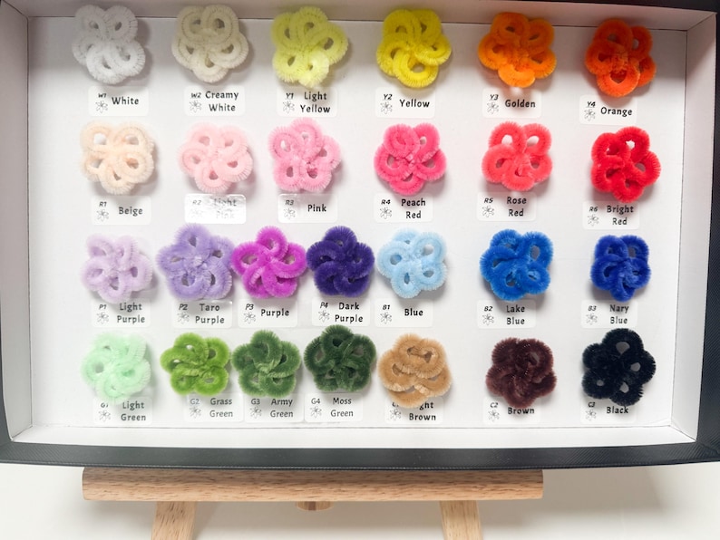 May include: A display of colorful, fluffy, flower-shaped craft supplies. The assortment includes white, yellow, orange, pink, red, purple, blue, green, brown, and black. Each color is labeled with a corresponding name.
