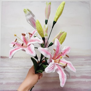 May include: A bouquet of handmade pink and white lily flowers with yellow stamens and green leaves. The lilies have a detailed, textured appearance, with pink stripes on the petals. The flower buds are a pale yellow-green colour.