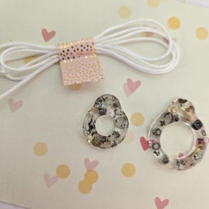 May include: Two clear resin pendants with embedded gears and metallic accents. The pendants are teardrop-shaped with a circular opening. A white cord and a pink tag with gold polka dots are also visible.