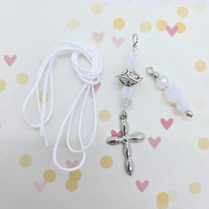 May include: A white rosary featuring a silver cross and decorative beads. The rosary includes a white cord and a separate strand of beads. The background is a light colour with pink hearts and yellow circles.