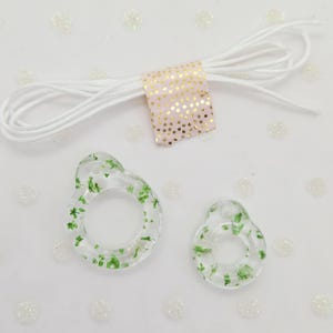 May include: Two clear, ring-shaped pendants with green floral inclusions, a white cord, and a pink ribbon with gold polka dots. The pendants are different sizes, possibly a jewellery or craft set.
