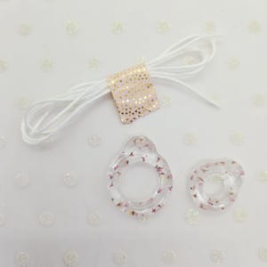 May include: Two clear resin pendants with pink flowers, one teardrop and one round. A white cord is bundled with a pink and gold polka dot ribbon. The pendants are on a white background with white polka dots.