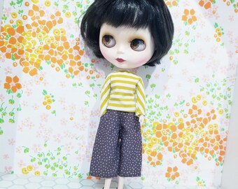 May include: A Blythe doll with black hair, wearing a yellow and white striped top and dark blue polka dot trousers. The doll is also wearing brown shoes with cat face details. The background features a floral pattern.