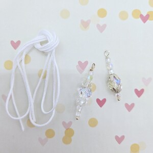 May include: Two white crystal beaded charms with silver accents and a white cord. The charms are on a white background with pink and yellow confetti.