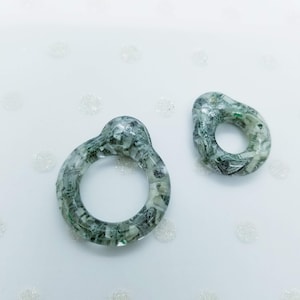 May include: Two resin rings, one with a loop and the other oval-shaped, filled with fragments of green and white currency. The rings are set against a white background with subtle, sparkling accents.