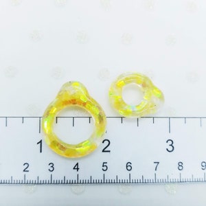 以下が含まれることがあります： 黄色と虹色のグリッターが埋め込まれた透明なレジン製のリングが2つ。リングの直径は約2.5cmと3.8cmです。