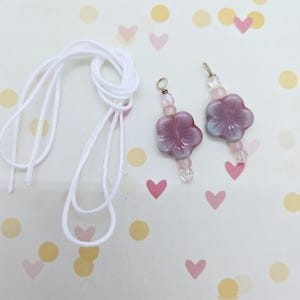 May include: Two pink flower charms with clear beads and a white cord. The charms are made of glass and have a delicate, feminine look.
