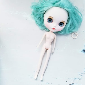 May include: A doll with a pale complexion, blue eyes, and turquoise hair. The doll has a slender body and is lying on a light blue surface. A small white accessory is next to the doll's head.