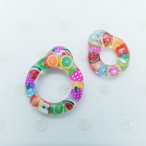 May include: Two clear resin rings decorated with a fruit theme. The rings feature colourful images of watermelon, strawberries, lemons, limes and kiwi.