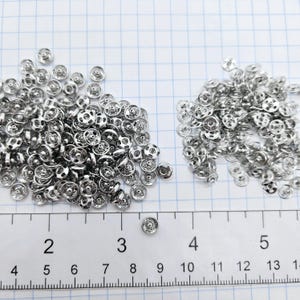 以下が含まれることがあります： 2つの山に配置された小さな円形の銀色の金属製スナップボタン部品の大きなコレクション。下部に定規が表示されており、個々の部品の直径が約0.6cmであることを示しています。