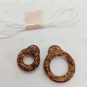 Peut inclure: Deux pendentifs en résine marron avec des paillettes dorées, en forme de breloques en trou de serrure. Un paquet de cordon élastique blanc est visible, maintenu par une bande rose et dorée. Fond blanc avec des flocons scintillants.