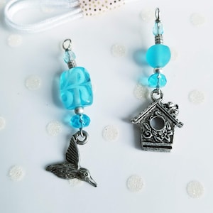 May include: Two handmade charm pendants. One features a hummingbird charm with blue glass beads. The other has a birdhouse charm with blue glass beads. Both charms have silver-coloured metal accents and are suspended from a white cord.