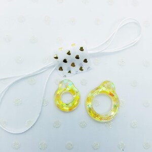 May include: Two yellow resin rings with gold glitter embedded in the resin. The rings are on a white background with small white dots.