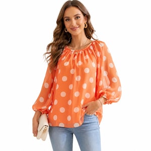 Puede incluir: Blusa naranja con lunares blancos. La blusa tiene mangas largas y abullonadas y un cuello redondo. La tela parece ligera y semitransparente. La blusa es de un color brillante y alegre y es adecuada para ropa informal.