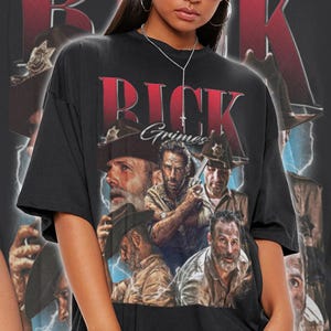 May include: Black unisex t-shirt featuring a graphic of Rick Grimes from The Walking Dead. The design includes the name "Rick Grimes" in red lettering and multiple images of the character. The shirt is oversized.