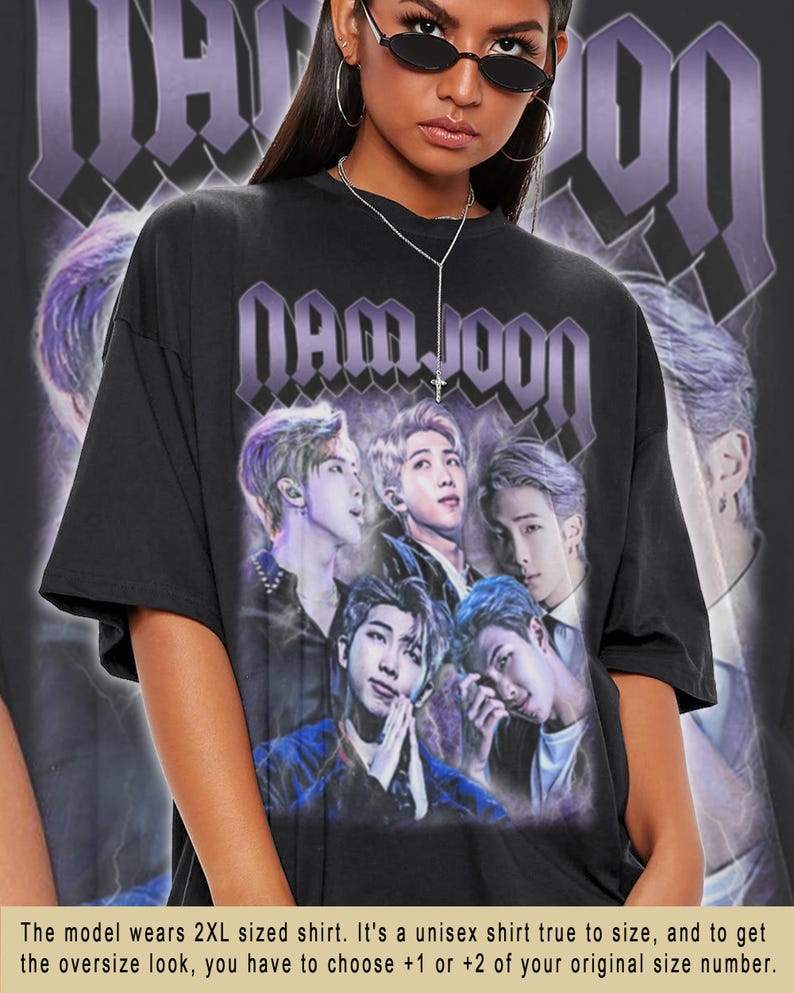 May include: Black t-shirt with a graphic print of a group of men with the text "NAMJOON" in purple letters. The model is wearing a 2XL size shirt.