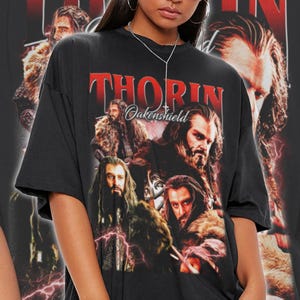 Może przedstawiać: Czarny, duży t-shirt z grafiką Thorina Oakenshielda i czerwonym napisem "THORIN Oakenshield". Projekt zawiera obrazy postaci w r&oacute;żnych pozach. Koszulka jest w stylu unisex.