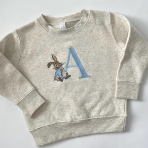 May include: A light gray sweatshirt featuring an embroidered Peter Rabbit design and the letter "A" in light blue. The rabbit is wearing a blue jacket. The sweatshirt has a crew neck and ribbed cuffs and hem.