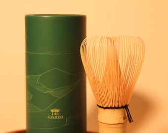 May include: A green cylindrical container with the text "GISHIKI" and a bamboo matcha whisk. The whisk has many thin tines and a bamboo handle. The container and whisk are on a wooden tray.