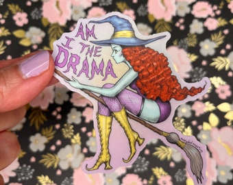 Peut inclure: Un autocollant représentant une sorcière de dessin animé aux cheveux roux, vêtue d'une tenue violette et noire et de bottes jaunes, chevauchant un balai. La sorcière regarde vers le bas et le texte "I am the drama" est écrit en lettres violettes sur un fond rose.