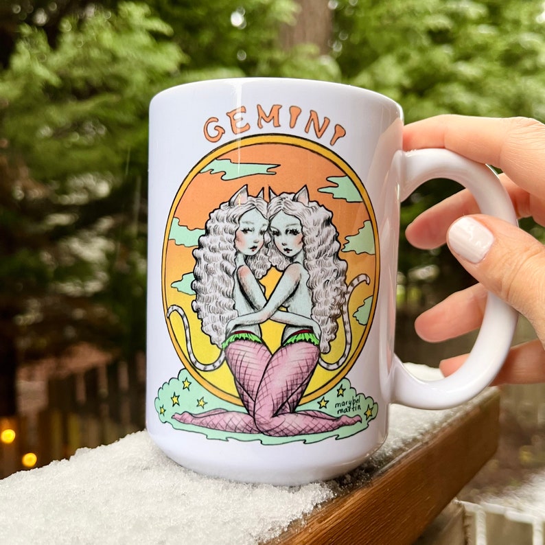 May include: White ceramic mug with a colorful illustration of two women with cat ears, holding each other. The illustration is surrounded by a circle with the word "GEMINI" at the top and a sunset background. The mug has a black handle and the artist's signature "marlyn martin" at the bottom.