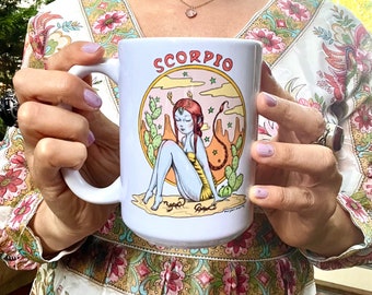 May include: White ceramic mug with a colorful illustration of a woman with a scorpion tail sitting on a desert landscape. The mug has the word "SCORPIO" printed in black letters.