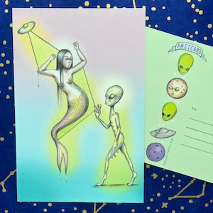 Könnte beinhalten: Eine skurrile Postkarte mit einer Meerjungfrau, die von einem UFO entführt wird, mit einem Alien, das zusieht. Die Illustration ist in Pastelltönen vor einem hellgrünen und rosa Hintergrund gehalten. Die Postkarte enthält auch ein kleineres Bild von Alien-Gesichtern und einem UFO.