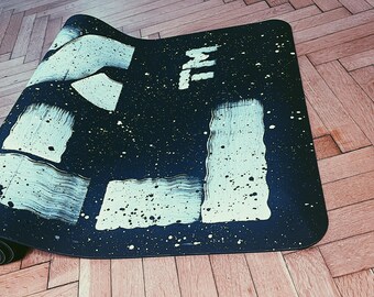 May include: A rolled-up black yoga mat with white brushstroke designs and the text "TM". The mat has white paint splatters and is on a wooden floor. The mat is designed for exercise and fitness.