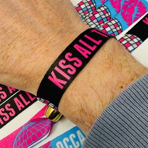 Może przedstawiać: Czarna opaska na nadgarstek z tkaniny z napisem "KISS ALL" w jaskrawym r&oacute;żu. Widoczne są dodatkowe opaski z grafiką kuli dyskotekowej i ust. Opaska jest zabezpieczona złotym zapięciem. Og&oacute;lny motyw związany jest z wydarzeniami lub imprezami.