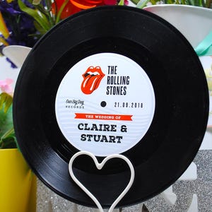 May include: A black vinyl record with a white label featuring the Rolling Stones logo and wedding details. The record is displayed on a white heart-shaped stand. The text on the record reads "The Wedding of Claire & Stuart".